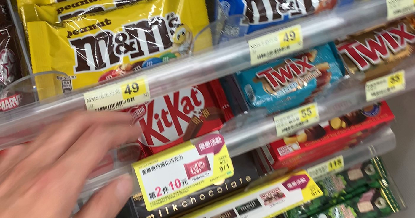 A 7-Eleven chocolate shelf in Taiwan with KitKat, M&M's, Twix and Snickers behind NT$ price tags, a hand reaching for a bar