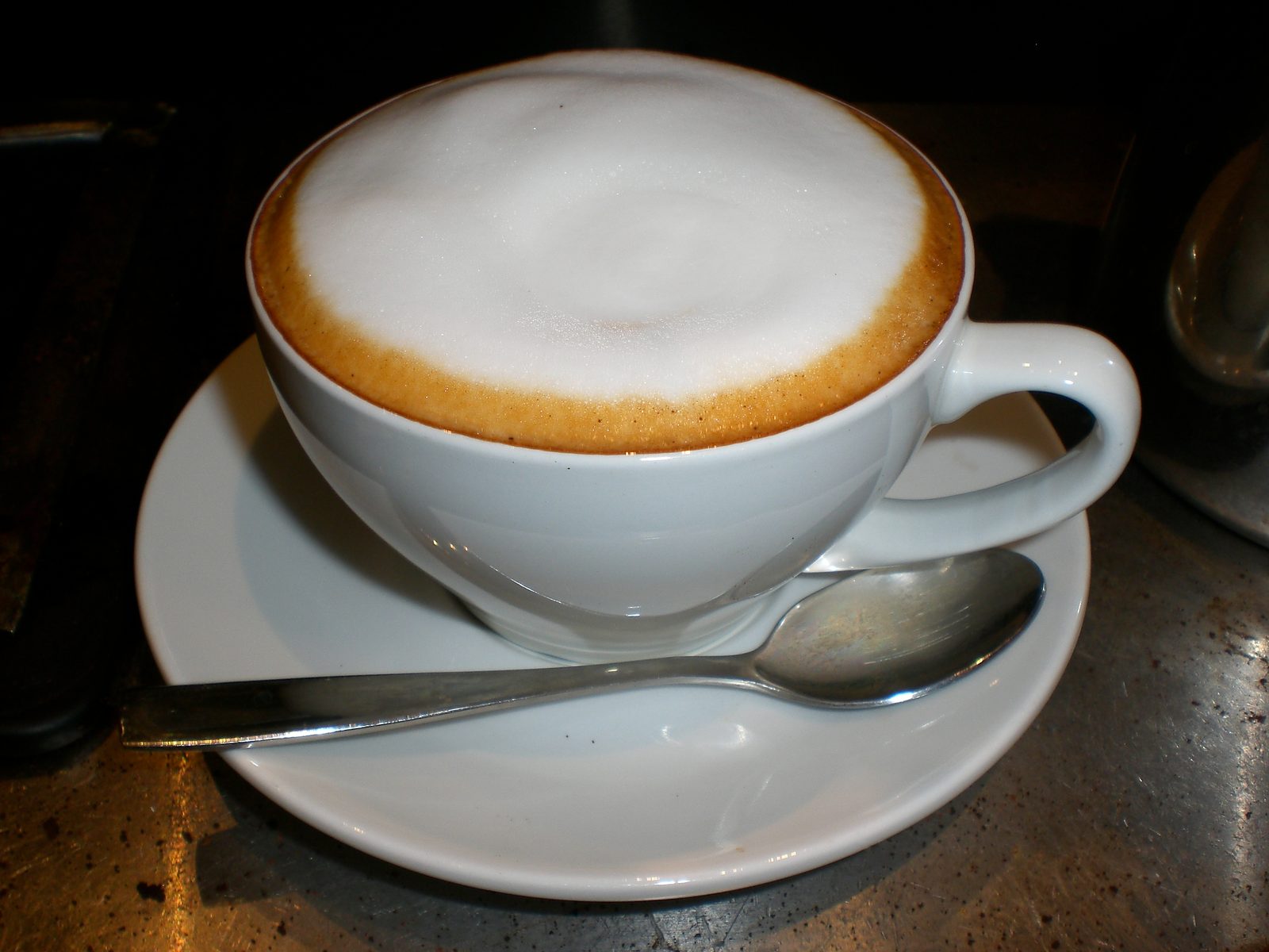 A white cappuccino cup full of milk foam on a saucer, with a spoon in front