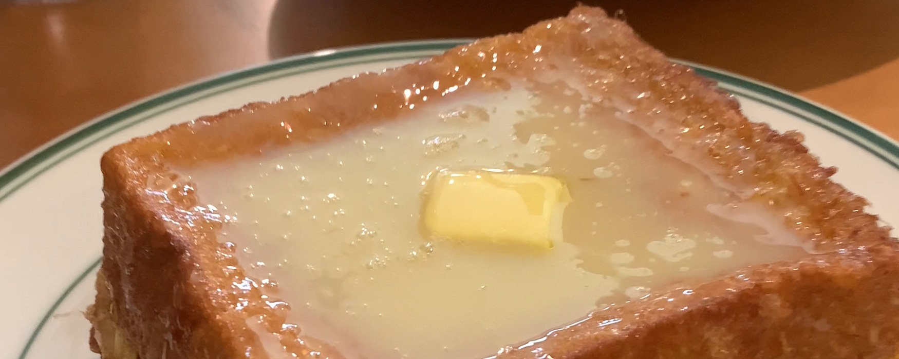 A Hong Kong-style french toast, deep-fried and golden, with a pat of butter melting into a pool of condensed milk, on a green-rimmed plate on a wooden cha chaan teng table