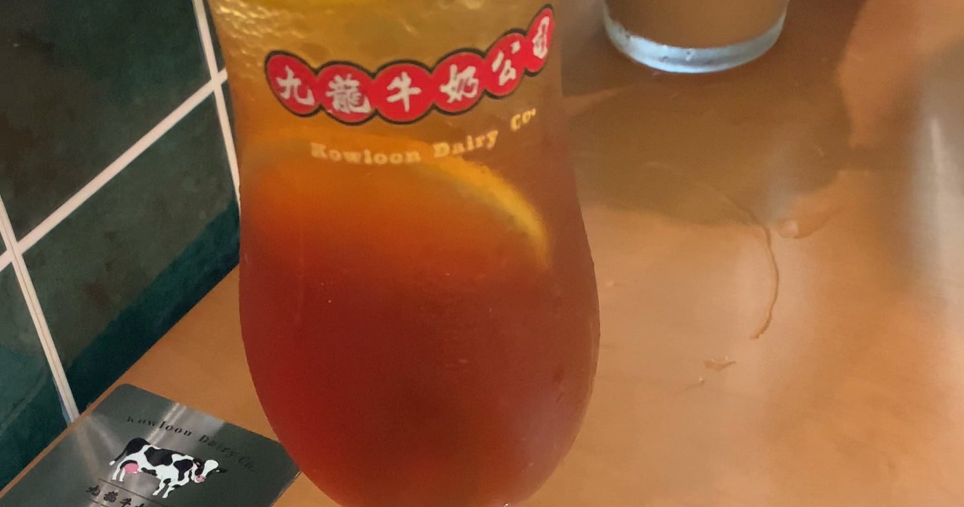 茶餐廳木桌上一杯港式凍檸茶，杯裡有檸檬片，背景是綠色磁磚牆