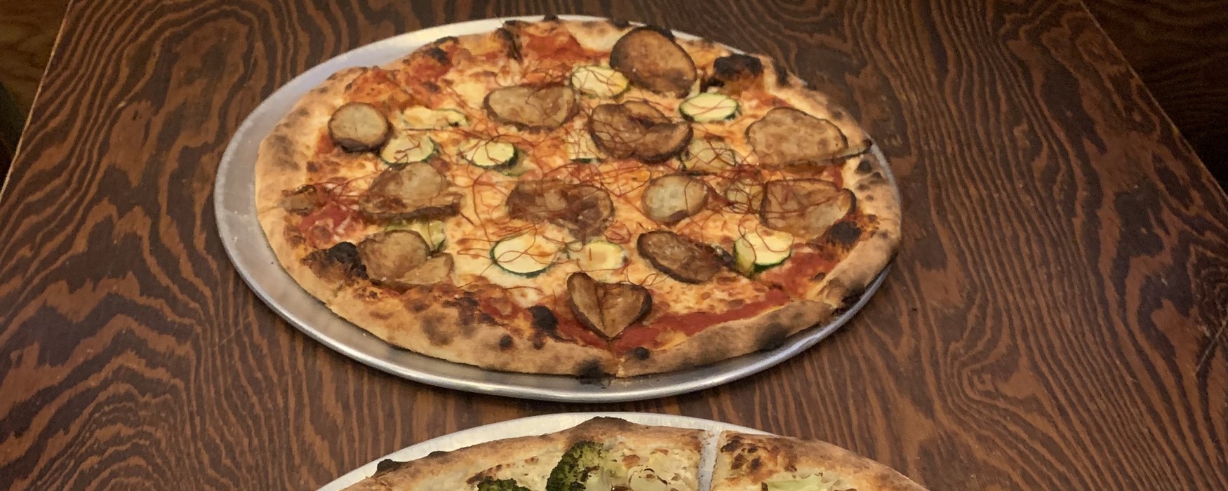 Two whole pizzas on metal trays on a dark wood table — an eggplant and zucchini pizza in front, a broccoli white pizza arriving behind it